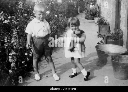 Un ragazzo e una ragazza in un giardino che si tengono per mano, la ragazza che stringe un orsacchiotto, Surrey, c1930. Si tratta di una fotografia tratta da un album di scatti, durante l'occupazione della Palestina da parte dell'esercito britannico, con foto provenienti dalla Palestina e dal Regno Unito, tra cui devil's Jump vicino a Churt nel Surrey e devil's Punch Bowl. L'addestramento sembra essere a Hursley Camp, vicino a Winchester. La Gran Bretagna amministrò la Palestina per conto della società delle Nazioni tra il 1920 e il 1948, un periodo denominato "mandato britannico”. Foto Stock