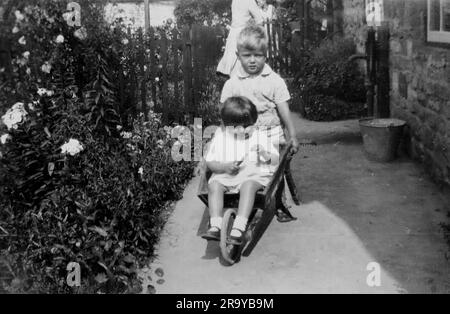 Un ragazzo e una ragazza in un giardino, seduto in una ruota di legno barrow mentre il ragazzo la spinge, Surrey, c1930. Si tratta di una fotografia tratta da un album di scatti, durante l'occupazione della Palestina da parte dell'esercito britannico, con foto provenienti dalla Palestina e dal Regno Unito, tra cui devil's Jump vicino a Churt nel Surrey e devil's Punch Bowl. L'addestramento sembra essere a Hursley Camp, vicino a Winchester. La Gran Bretagna amministrò la Palestina per conto della società delle Nazioni tra il 1920 e il 1948, un periodo denominato "mandato britannico”. Foto Stock