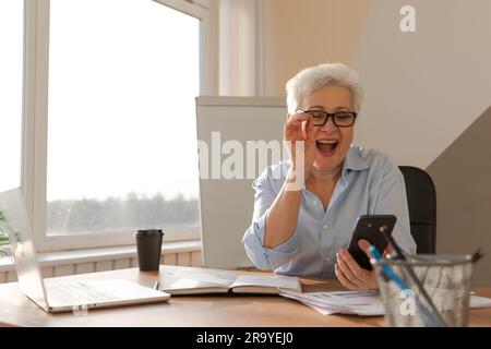 Sicura donna anziana di mezza età euforica vincitrice con smartphone. Signora più anziana matura guardando il telefono delle cellule che legge le notizie grandi ottenendo il buon risultato che vince la sensibilità in linea dell'offerta stupita. Gesto vincente Foto Stock