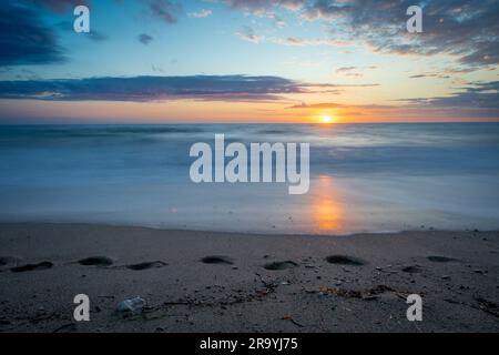 Lunga esposizione di impronte e onde che si gettano su una sabbia durante il tramonto. Foto Stock