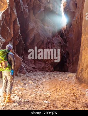 Un escursionista, in piedi, che guarda la luce che passa attraverso una stretta apertura nelle pareti di un canyon a fessura illuminata da luci soffuse riflettenti, Foto Stock