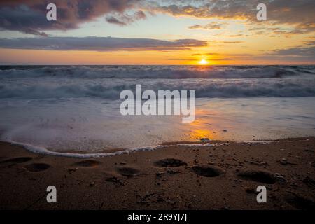 Lunga esposizione di impronte e onde che si gettano su una sabbia durante il tramonto. Foto Stock