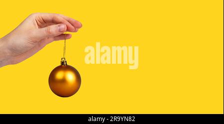 Palla di Natale marrone oro, bauble, ornamento su corda, appeso a mano femminile. Striscione monocromatico giallo. Foto Stock