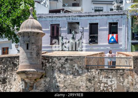 Una donna che ammira la vista dal vecchio muro della fortezza di Old San Juan Puerto Rico con sculture e una bandiera portoricana dietro di lei. Foto Stock