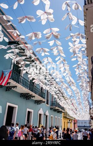 Calle Fortaleza (ex strada degli ombrelli) nella Vecchia San Juan, Porto Rico, con rastrellatori bianchi di farfalle sopra la testa e bandiera portoricana sopra gli acquirenti. Foto Stock