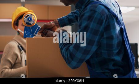 Uomo afro-americano che applica nastro adesivo sulle scatole, prepara l'ordine con merce da magazzino in magazzino. Dipendente maschio che sigilla i pacchi con i prodotti, servizio di consegna. Foto Stock