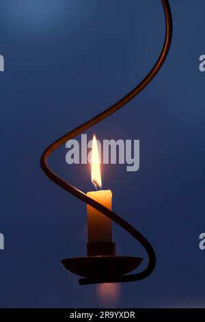 Primo piano interno di una candela accesa su un portacandele ruotato appeso in una finestra su sfondo blu al crepuscolo all'esterno Foto Stock