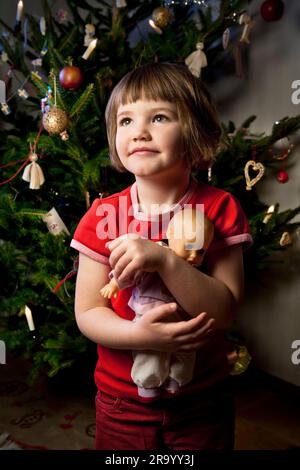 Bambina con la bambola che guarda lontano nella parte anteriore di un albero di Natale Foto Stock