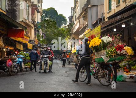 Un venditore di fiori spinge la sua bicicletta carica oltre i negozi di Cau Go, all'interno del quartiere vecchio di Hanoi, in Vietnam. Foto Stock