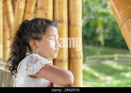 Piccola ragazza latina con pelle marrone, molto seria e annoiata, pensando al bullismo che riceve, in piedi sul ponte di bambù. Foto Stock