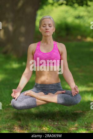 Una giovane donna con i capelli biondi, che indossa un top raccolto, siede a gambe incrociate in posizione Lotus, levita sopra l'erba, fa yoga, meditando pacificamente. Foto Stock