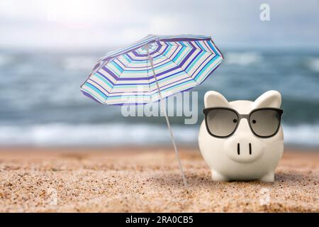 Sconti per le vacanze. Salvadanaio con occhiali da sole e ombrellone sulla spiaggia sabbiosa vicino al mare Foto Stock