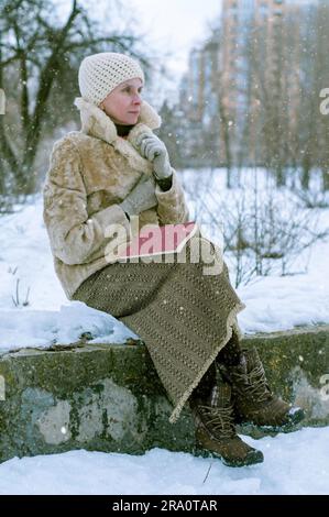 Una donna adulta con un cappotto di pelliccia e un berretto di lana è seduta su una panchina di pietra e sta leggendo un libro in inverno, in un parco coperto dalla neve Foto Stock