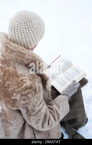 Una donna adulta con un cappotto di pelliccia e un berretto di lana è seduta su una panchina di pietra e sta leggendo un libro in inverno, in un parco coperto dalla neve Foto Stock