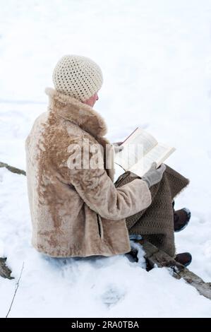 Una donna adulta con un cappotto di pelliccia e un berretto di lana è seduta su una panchina di pietra e sta leggendo un libro in inverno, in un parco coperto dalla neve Foto Stock