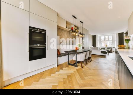una cucina moderna con pavimento in legno e armadi bianchi al centro della sala è un forno, tavolo da pranzo e sedie Foto Stock
