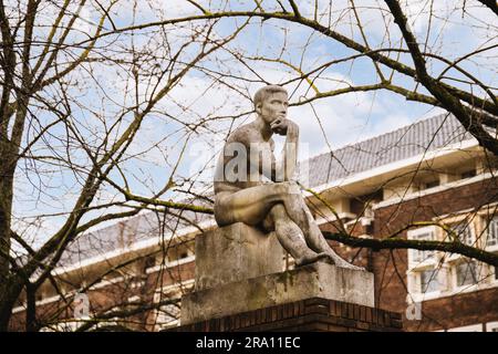 una statua seduta in cima ad un blocco di pietra di fronte ad un edificio in mattoni con alberi e cielo sullo sfondo Foto Stock