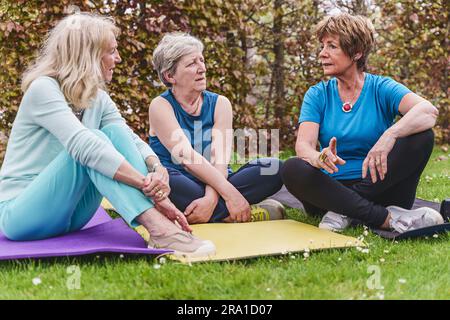 Tre donne anziane siedono sui tappetini yoga all'aperto sull'erba, chiacchierando insieme dopo una sessione di allenamento. Foto Stock