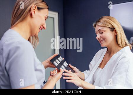 La giovane donna bionda in un cappotto bianco sceglie gli orecchini per la procedura di perforazione dell'orecchio in un salone di bellezza. Una consulente sorridente in uniforme grigia offre un Foto Stock