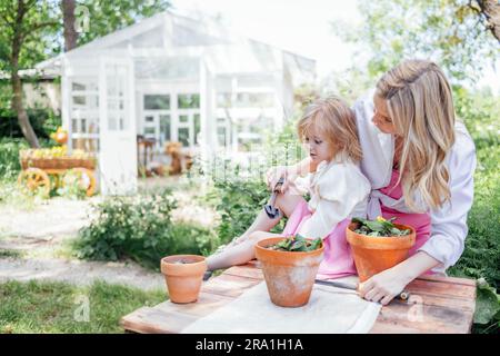 La bambina e la sua bella mamma bionda stanno piantando fiori in vasi di argilla all'aperto in giardino. La giovane donna e sua figlia carina passano del tempo insieme in b Foto Stock