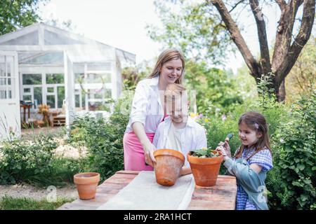 La giovane donna e i suoi due figli stanno piantando fiori in vasi di argilla in giardino. Una madre attraente, una ragazza carina e un ragazzo nel cortile. Un felice Foto Stock
