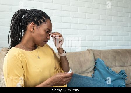 Donna malata che controlla il risultato del termometro. Donna afro-americana in affari a casa per misurare la temperatura corporea con il termometro, sentendosi malata Foto Stock