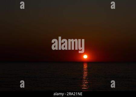 Un grande sole arancione che tramonta su un mare calmo. Scatto statico scattato durante una serata senza nuvole. Immagine sotto esposta nello scopo Foto Stock