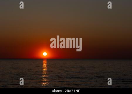 Un grande sole arancione che tramonta su un mare calmo. Scatto statico scattato durante una serata senza nuvole. Immagine sotto esposta nello scopo Foto Stock