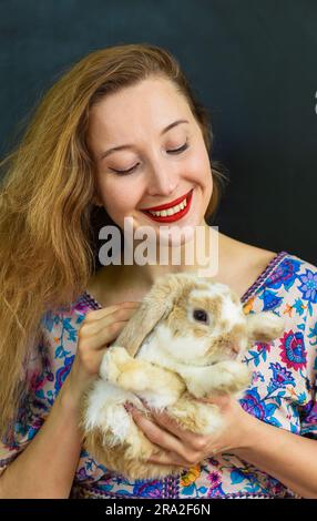 Bella donna russa con lunghi capelli biondi che tiene in mano un animale domestico di coniglio contro il muro nero Foto Stock