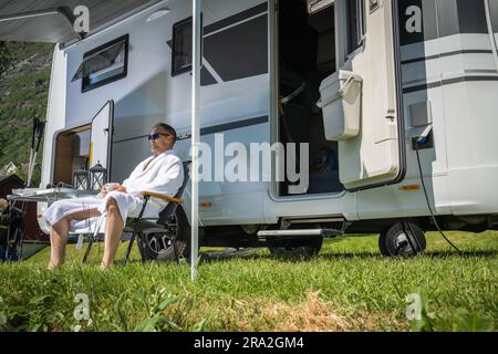 Un uomo caucasico che indossa un accappatoio che si gode la mattina presto in un parcheggio per camper di fronte al suo camper Van. Tema viaggi e attività all'aperto. Foto Stock