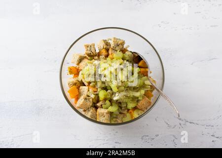 Una ciotola di vetro con una miscela di cubetti di zucca, crostini, sedano stufato con cipolle e spezie su sfondo azzurro, vista dall'alto. Fase di cottura de Foto Stock