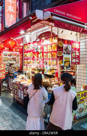 Due giovani donne giapponesi che guardano una bancarella cinese di cibo da asporto che vende panini e gnocchi al vapore, la notte a Nankinmachi, Chinatown, Kobe. Foto Stock