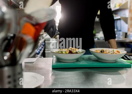 Coltivate uno chef anonimo maschio in uniforme nera che serve una deliziosa insalata con formaggio alla griglia in cucina del ristorante Foto Stock