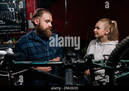 Ritratto di un riparatore di ciclismo che parla con una cliente bionda, parlando di un problema di bicicletta rilevato durante la diagnostica in riparazione Foto Stock