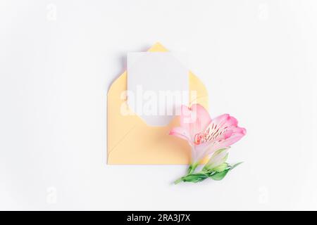Busta gialla con scheda vuota e fiore di alstroemeria rosa su sfondo bianco. Compleanno, San Valentino, festa della mamma, concetto di giorno delle donne. Vista dall'alto, Foto Stock