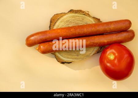 due salsicce si trovano e un pomodoro rosso su una tavola rotonda di legno su fondo marrone da vicino Foto Stock