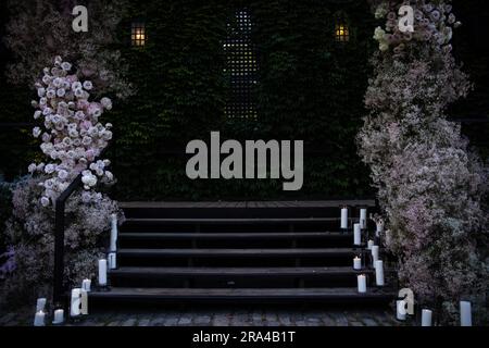 Bellissime composizioni di fiori arrossati per matrimoni con rose, candele e fiato del bambino in un lussureggiante evento con giardino botanico con scale e arco. Foto Stock