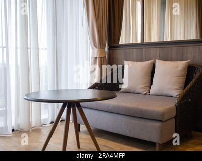 Vuoto, moderno e accogliente divano marrone, realizzato in vimini e cuscino grigio con due cuscini, tavolino rotondo vuoto con pavimento in legno vicino al vetro sli Foto Stock