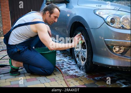 Bell'uomo caucasico giovane adulto, operaio di servizio auto in tuta blu, lavaggio manuale dei cerchi in lega di auto con schiuma detergente e spugna. Il conce Foto Stock