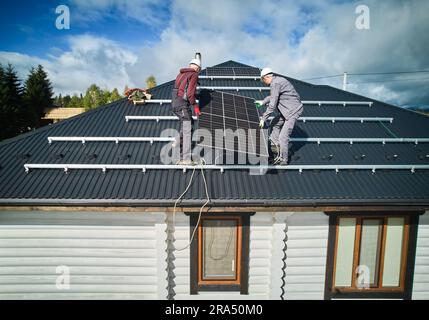 Lavoratori maschi che installano celle solari su un tetto. Installazione del sistema solare sulla parte superiore della casa. Due uomini tecnici che trasportano un pesante pannello solare su un tetto nelle giornate di sole. Foto Stock