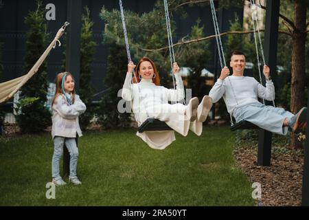 Mamma e papà stanno cavalcando su un'oscillazione e ci è una figlia in piedi accanto a loro. La famiglia sta riposando su un'oscillazione. Foto Stock