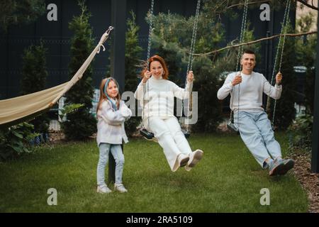 Mamma e papà stanno cavalcando su un'oscillazione e ci è una figlia in piedi accanto a loro. La famiglia sta riposando su un'oscillazione. Foto Stock