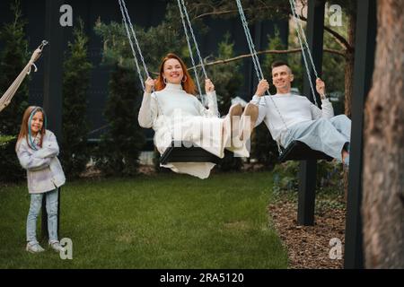 Mamma e papà stanno cavalcando su un'oscillazione e ci è una figlia in piedi accanto a loro. La famiglia sta riposando su un'oscillazione. Foto Stock