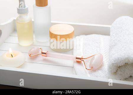 Strumento per la laminazione del viso in pietra di quarzo rosa, vari prodotti generici per la cura del viso e del corpo creme idratanti oli su vassoio di legno bianco Luce studio. Foto Stock