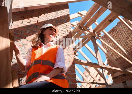 Lavoratore edile donna in piedi su una scala in legno con giubbotto di sicurezza e elmetto Foto Stock