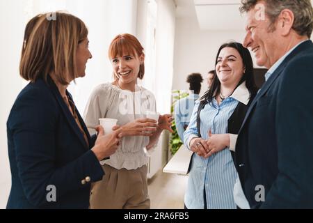 Gruppo di colleghi in abbigliamento formale conversando e ridendo vicino a una finestra durante una pausa di lavoro. Mix di senior e junior, in un ufficio moderno. Foto Stock