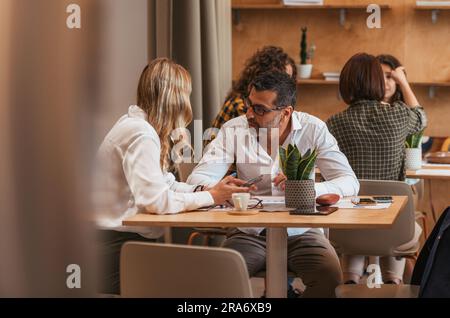 Giovane donna d'affari bionda che mostra all'uomo d'affari anziano qualcosa sul suo telefono al bar Foto Stock