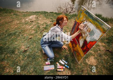 Ragazza con i capelli rossi in un panino che dipinge su una grande tela mentre si siede sull'erba Foto Stock