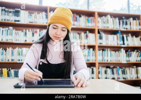 Attività accademiche: Vista frontale della giovane studentessa universitaria femminile che studia con il suo tablet e la sua penna in biblioteca Foto Stock