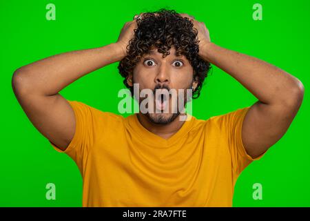 Oh mio Dio, wow. L'uomo vincitore entusiasta che alza le mani di sorpresa guardando la fotocamera con gli occhi grandi sconvolti dalla vittoria improvvisa. Sincero ragazzo indiano bello isolato da solo su sfondo cromatico verde Foto Stock